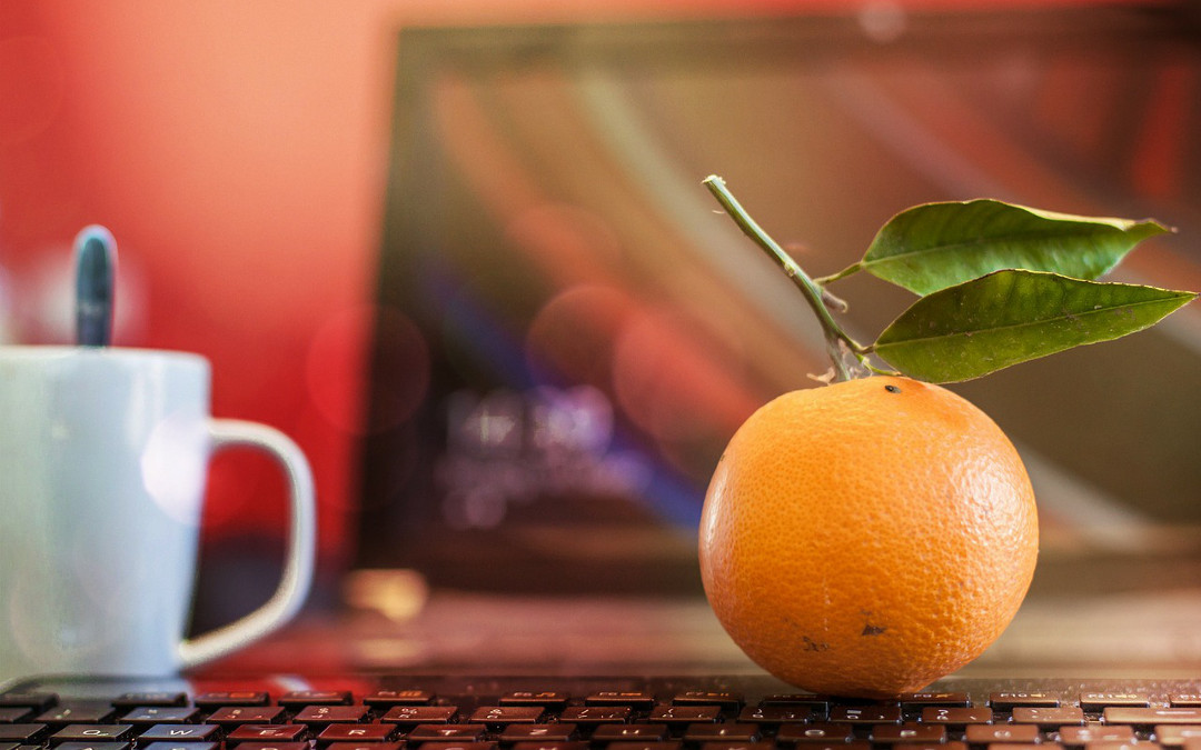 Eine Orange mit Blatt liegt auf einer Computertastatur. Verschwommen links im Hintergrund: ein Kaffeebecher.